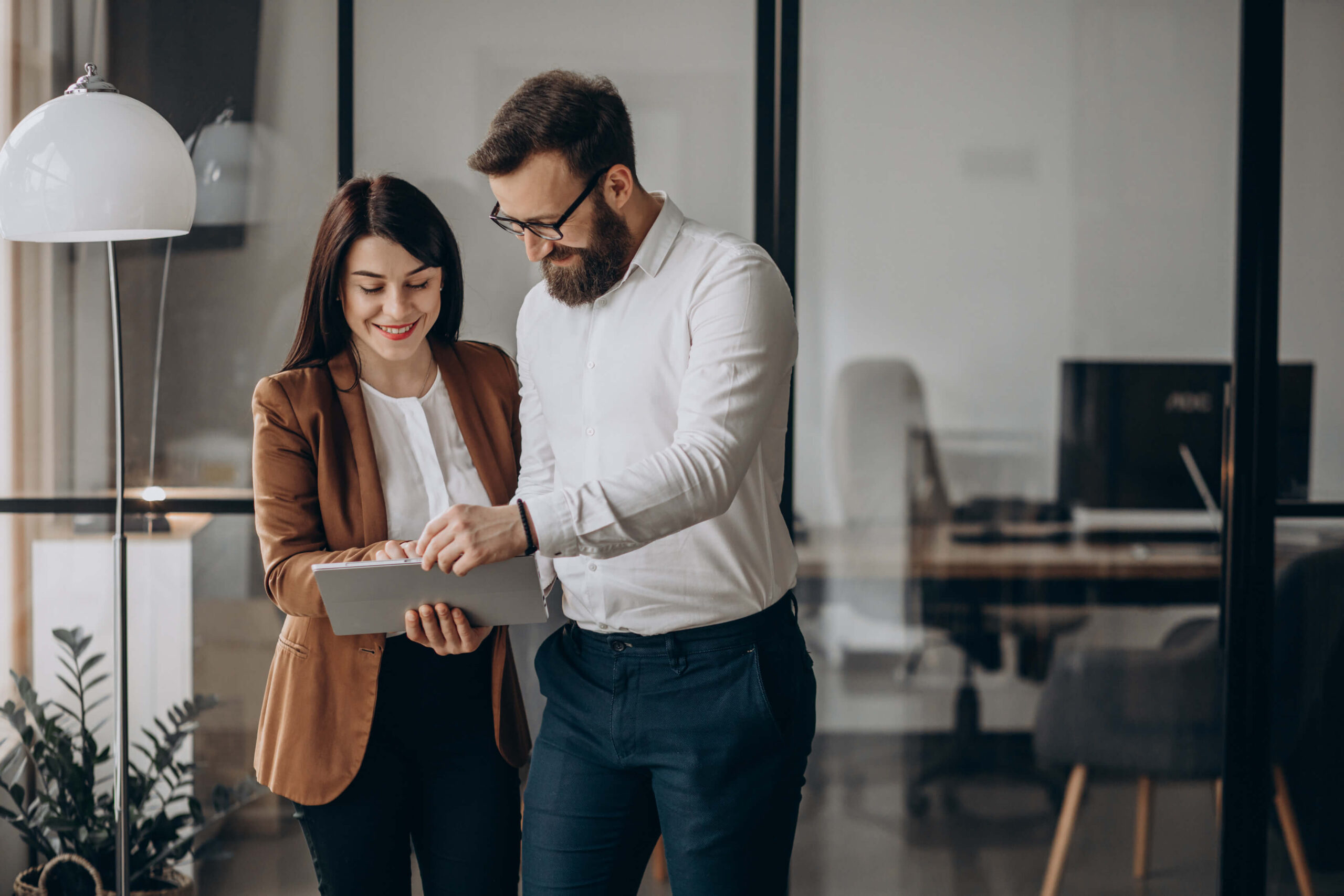 Two business partners in an office working together on a tablet, both smiling and reviewing business financing solutions in a modern, professional workspace with glass walls